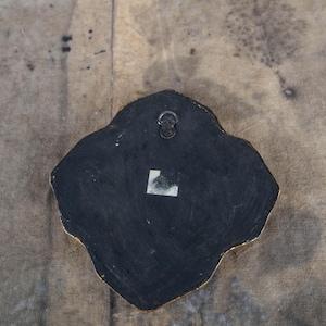 May include: Back of a decorative wall hanging with a black surface and a gold-colored edge. A metal hanger is attached at the top. A small white sticker is affixed to the lower portion of the black surface. The shape is irregular, with rounded edges.