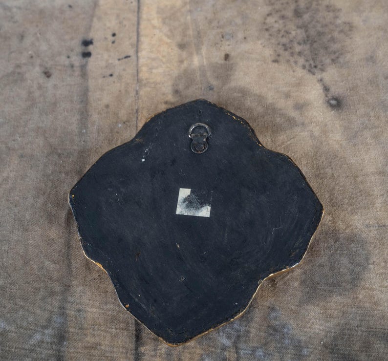 May include: Back of a decorative wall hanging with a black surface and a gold-colored edge. A metal hanger is attached at the top. A small white sticker is affixed to the lower portion of the black surface. The shape is irregular, with rounded edges.