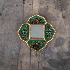 May include: A small, decorative mirror with a square center and a unique, four-lobed shape. The frame is gold with a green border, adorned with floral designs in red, white, and yellow. The mirror is set against a wooden surface.