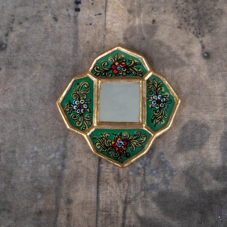 May include: A small, decorative mirror with a square center and a unique, four-lobed shape. The frame is gold with a green border, adorned with floral designs in red, white, and yellow. The mirror is set against a wooden surface.