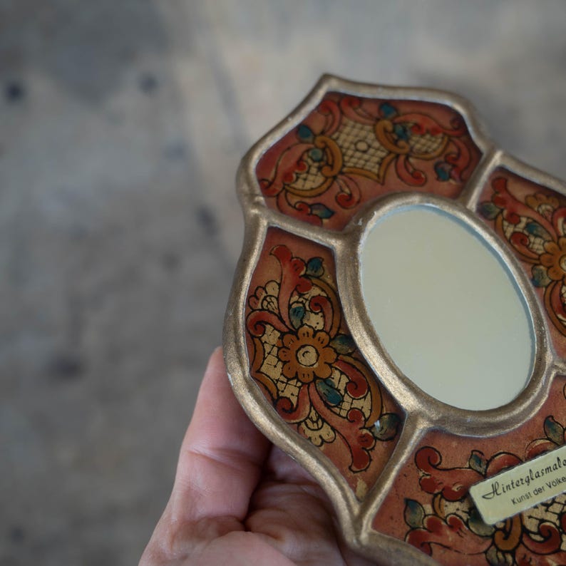 May include: Ornate, hand-painted mirror with a gold-colored frame. The mirror features a central oval surrounded by a decorative, floral design in red, gold, and teal. The mirror is held in a hand.