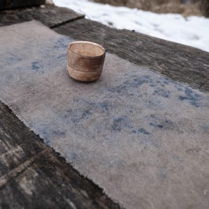 May include: A small, handmade, beige ceramic cup with a rough texture sits on a gray and blue textile. The cup has a simple, cylindrical shape with a slightly flared rim. The textile has a frayed edge and a mottled pattern.