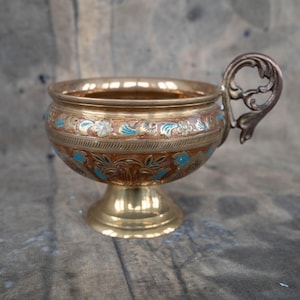 May include: A brass-colored decorative bowl with a pedestal base and ornate handle. The bowl features a floral design with blue and white accents on a brown background. The rim and base are polished brass.
