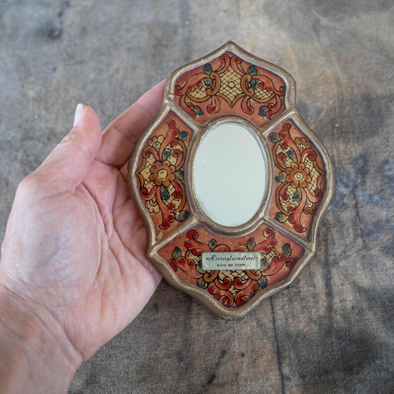 Ornate, vintage-style mirror with a gold-colored frame. The mirror features a decorative design in shades of red, orange, green, and yellow. The text "Hinterglasmalerei Kunst der Volker" is visible on a small label.
