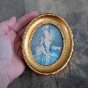 An oval miniature portrait held in a person's hand, displaying the scale and gold frame finish.