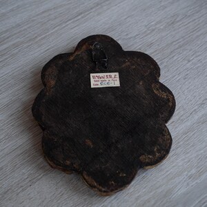 May include: Back of a dark brown, flower-shaped wooden wall hanging with a metal hanger. A small white label reads "U. Yuri S.R.L. hand made in Peru Code ECR-1."