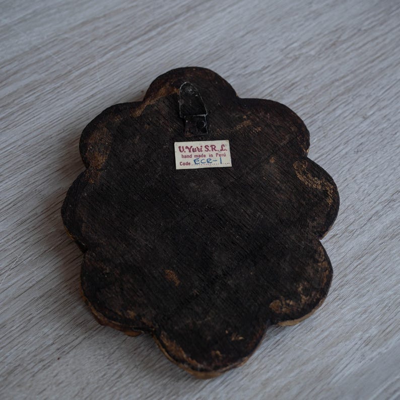 May include: Back of a dark brown, flower-shaped wooden wall hanging with a metal hanger. A small white label reads "U. Yuri S.R.L. hand made in Peru Code ECR-1."