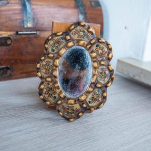 May include: A small, ornate, oval-shaped mirror with a gold-colored frame. The frame features a floral design with red, yellow, and green accents. The mirror is mounted on a wooden stand, and the background includes a wooden chest.