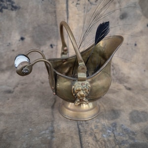 May include: A vintage brass coal scuttle with a handle and a decorative lion's head. The scuttle features a white and black ceramic handle and is filled with black feathers. The scuttle is approximately 6 inches tall.