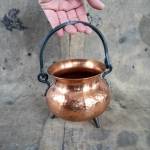 Peut inclure: Un petit chaudron en cuivre martelé avec une poignée en métal noir et trois petits pieds. Le chaudron a un corps rond et une surface brillante et réfléchissante. La poignée est fixée au chaudron avec des éléments métalliques décoratifs. Le chaudron est tenu par une main.