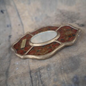 May include: An ornate, oval-shaped mirror with a red and gold decorative frame. The frame features floral patterns in red, gold, and blue. The mirror is small and has a reflective surface. The item is likely a decorative piece.