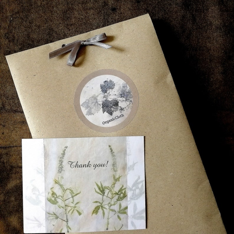 May include: A kraft paper package tied with a silver ribbon, featuring a round sticker with a botanical design and the text "OrganicCloth." A small card with a floral print and the words "Thank you!" is also included. The overall aesthetic is rustic and natural.