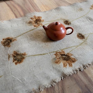 Peut inclure: Une théière en céramique brun rougeâtre de forme ronde est posée sur un chemin de table rectangulaire en lin beige clair. Le chemin présente un bord effiloché et un motif botanique de fleurs brunes reliées par de fines tiges courbes vert clair.