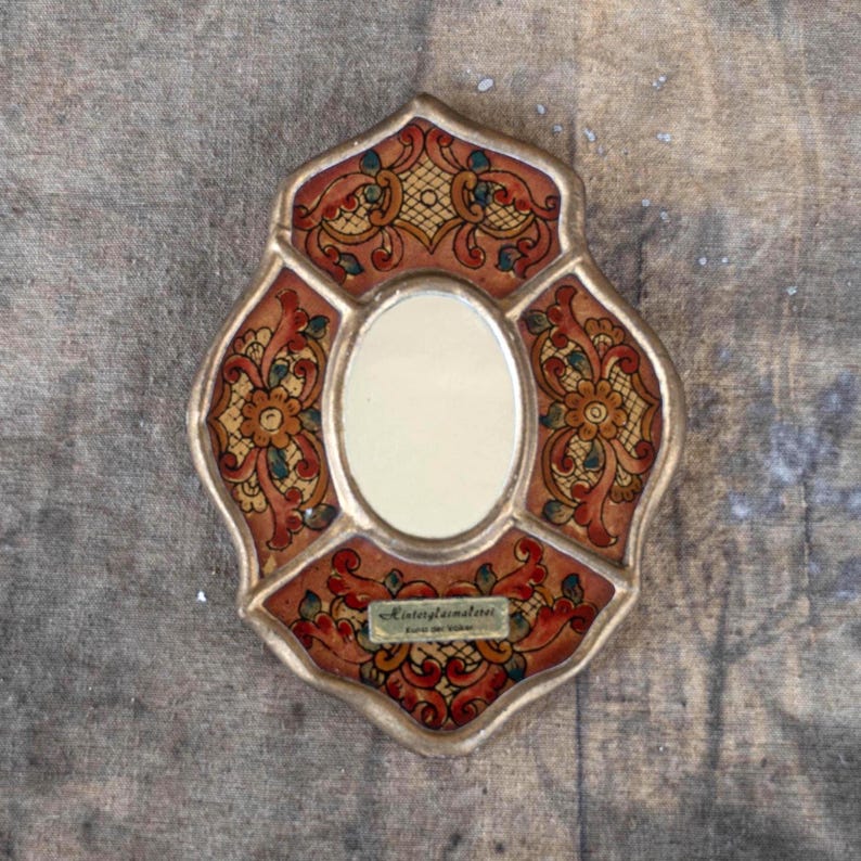 Vintage small decorative mirror oval shaped with reverse glass painting, floral design. The mirror has a frame with gold border, gold and brown colored