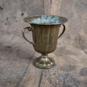 May include: A vintage brass cup with two handles and a fluted body. The cup has a decorative base and a slightly flared rim. The brass has a patina, with some areas showing a greenish hue, suggesting age and use.