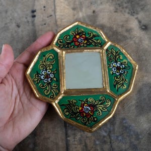 May include: A small, decorative mirror with a gold-painted frame. The frame has a green background with floral designs in red, white, and yellow. The mirror is square-shaped and set within an octagonal frame.