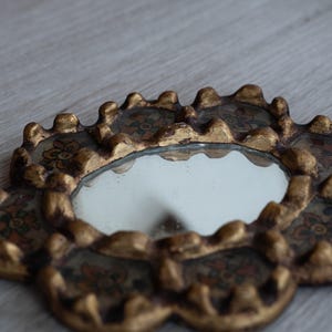 May include: A small, ornate mirror with a gold-colored, scalloped frame. The frame features floral designs in shades of red, brown, and green. The mirror itself is round and reflects the surrounding environment.