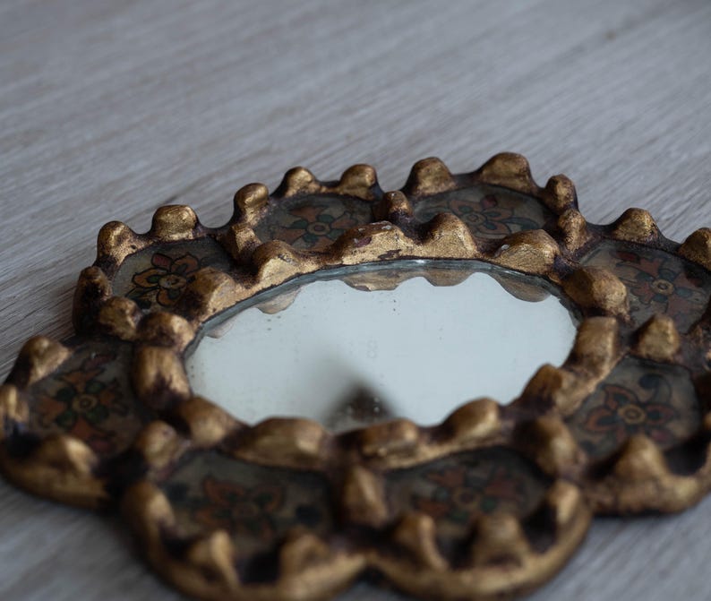 May include: A small, ornate mirror with a gold-colored, scalloped frame. The frame features floral designs in shades of red, brown, and green. The mirror itself is round and reflects the surrounding environment.