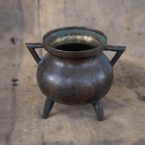 Peut inclure: Un petit chaudron antique en bronze avec trois pieds et deux poignées. Le pot a un corps arrondi et une large ouverture. L'intérieur du pot a une teinte dorée, tandis que l'extérieur présente une patine sombre et vieillie. Le chaudron est probablement utilisé à des fins décoratives.
