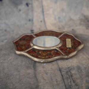 An ornate, small decorative mirror with a gold-colored frame and a central oval mirror. The frame features a red and gold floral pattern. A small label is affixed to the frame.