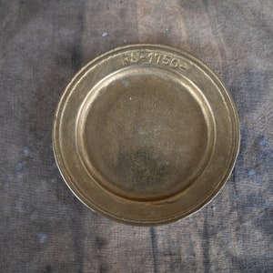 May include: A gold-coloured metal plate with a raised rim and a smooth surface. The plate has the inscription "KS-1150" around the rim.