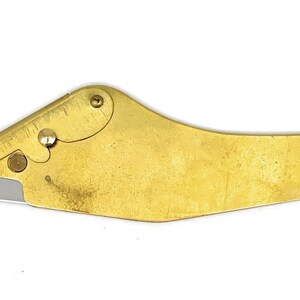 May include: A gold-colored, curved, handheld utility knife with a silver blade. The knife has a small, round hole at the end of the handle.