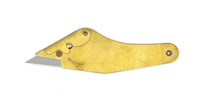 May include: A gold-colored, curved, handheld utility knife with a silver blade. The knife has a small, round hole at the end of the handle.