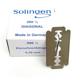 May include: A package of 200 1/2 Diagonal razor blades, made in Germany. The package is white with blue text and a blue logo. The razor blade is silver and has a diagonal cut. The text on the package reads "Solingen", "200 1/2 Diagonal", "Made in Germany", "K&uuml;rschnerklinge", and "0,20 mm".