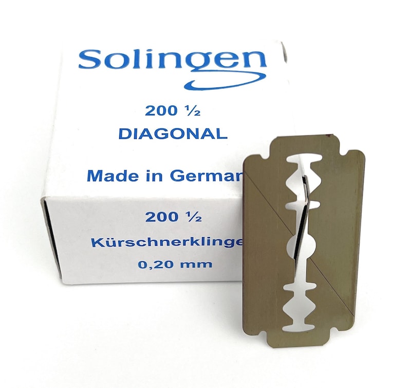 May include: A package of 200 1/2 Diagonal razor blades, made in Germany. The package is white with blue text and a blue logo. The razor blade is silver and has a diagonal cut. The text on the package reads "Solingen", "200 1/2 Diagonal", "Made in Germany", "K&uuml;rschnerklinge", and "0,20 mm".