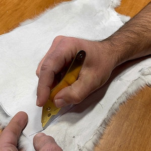 May include: A person's hand using a small gold tool to cut a piece of white fur. The fur is lying on a wooden surface.