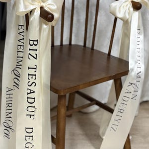 Op de afbeelding: Een houten stoel met een gebogen rugleuning en armleuningen. Crèmekleurige linten met strikken zijn aan de armen van de stoel vastgemaakt. De linten hebben zwarte tekst in een vreemde taal. De stoel staat op een lichte vloer.