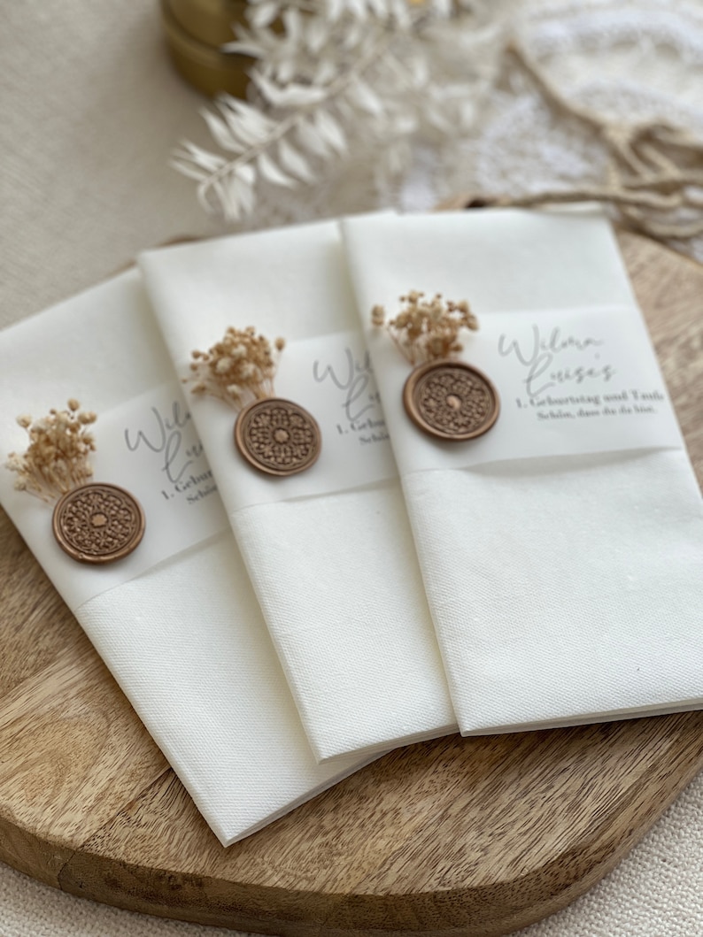 May include: Three white folded cards with a brown wax seal and dried flowers on top. The cards have the text "Wilma Louise" and "1. Geburtstag und Taufe" written on them.