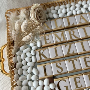 May include: A gold tray with white sugared almonds, a white rose, and dried flowers. The tray has a gold border and the words "ALLAH EMRİ KIZİ İSTE GEL" are spelled out in gold letters on white squares.