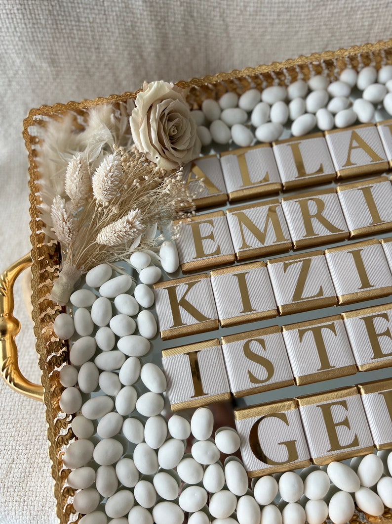May include: A gold tray with white sugared almonds, a white rose, and dried flowers. The tray has a gold border and the words "ALLAH EMRİ KIZİ İSTE GEL" are spelled out in gold letters on white squares.