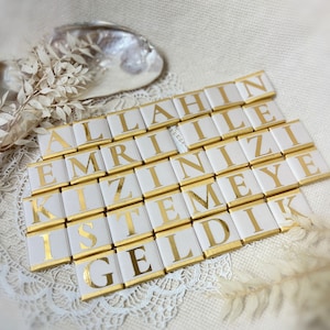 May include: White chocolate bars with gold foil wrappers, each bar has a letter printed on it, spelling out the phrase "ALLAHIN EMRINIZE KIZIMIZE ISTEMEYE GELDIK".
