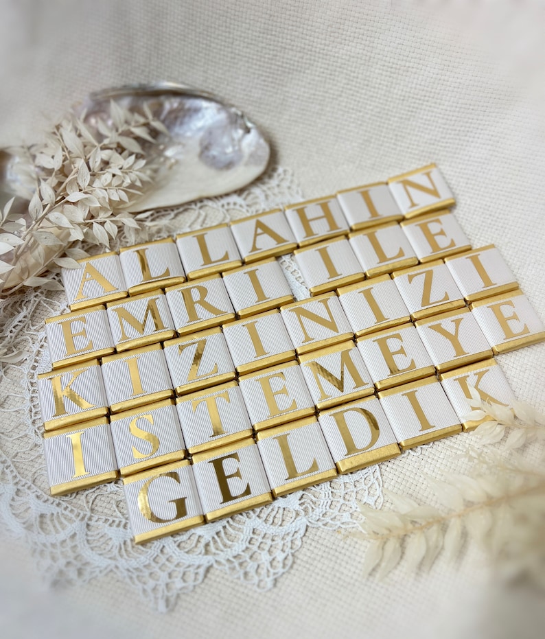 May include: White chocolate bars with gold foil wrappers, each bar has a letter printed on it, spelling out the phrase "ALLAHIN EMRINIZE KIZIMIZE ISTEMEYE GELDIK".