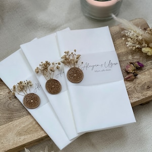 May include: Three white envelopes with a wax seal and dried flowers. The envelopes are stacked on top of each other. The top envelope has the text "Aleyna & Ugur" and "Alper, 24th June" written on it.