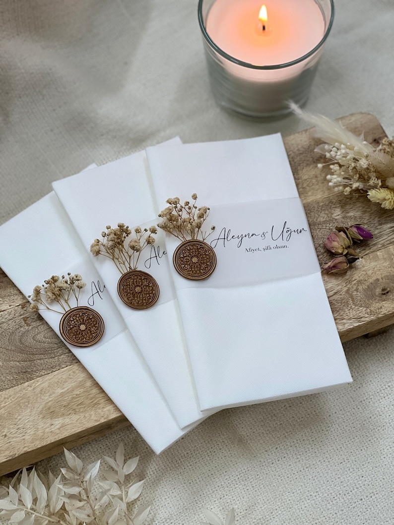 May include: Three white envelopes with a wax seal and dried flowers. The envelopes are stacked on top of each other. The top envelope has the text "Aleyna & Ugur" and "Alper, 24th June" written on it.