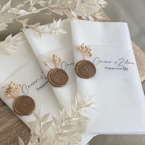 May include: Three white envelopes with gold wax seals and dried flowers. The envelopes are labeled with the text "Canen & Zilan Engagement".