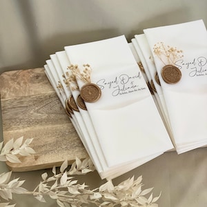 May include: A stack of white napkins with a gold wax seal and dried flowers. The napkins have the text "Sayed David & Juliana" and "Sch&ouml;n, dass du da bist." printed on them.