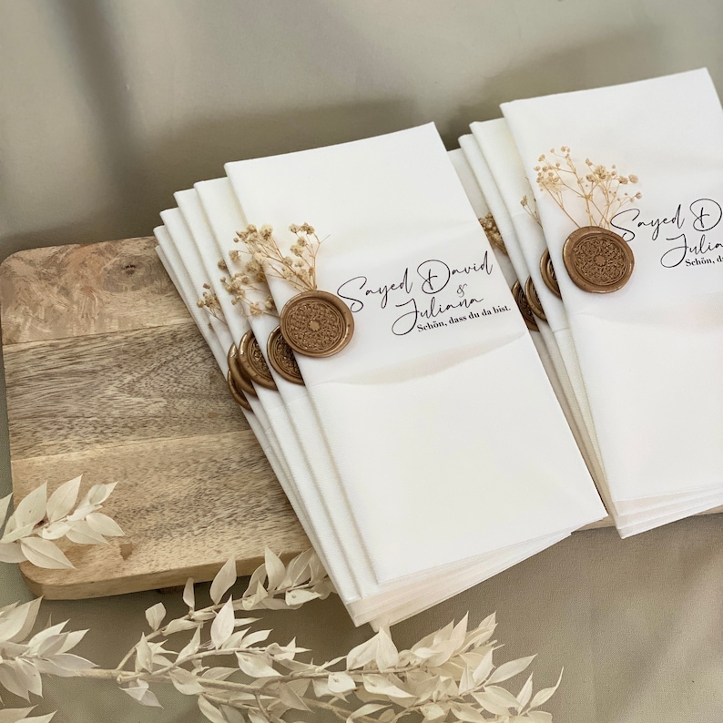 May include: A stack of white napkins with a gold wax seal and dried flowers. The napkins have the text "Sayed David & Juliana" and "Sch&ouml;n, dass du da bist." printed on them.