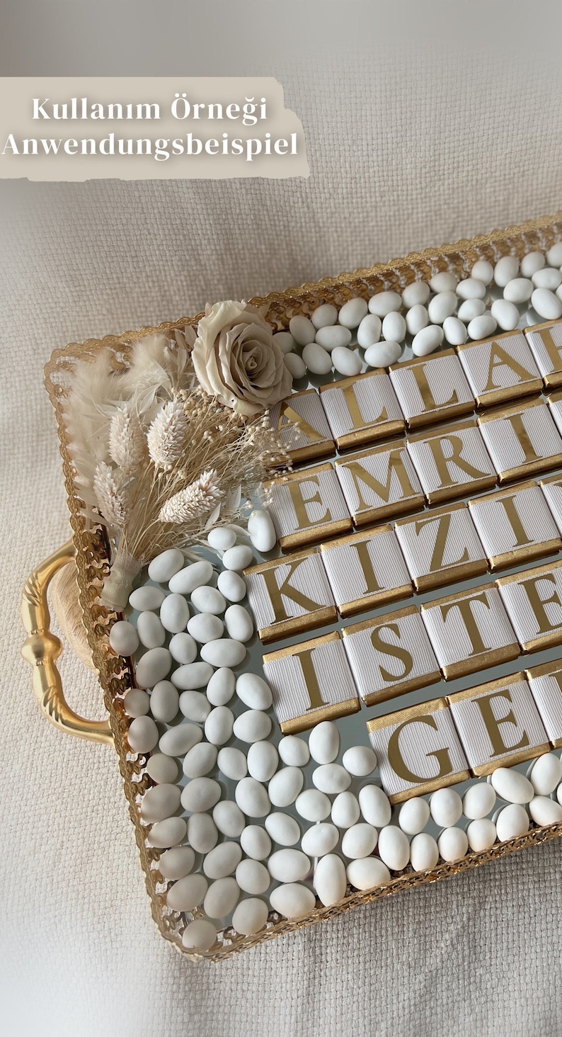 May include: A gold tray with white chocolate squares that spell out "ALLAH EMRİ KIZ İSTE GE", surrounded by white sugar almonds and dried flowers.