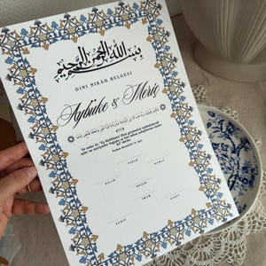 Pode incluir: Um certificado de casamento branco com uma borda floral azul, dourada e marrom ornamentada. O documento apresenta escrita árabe e os nomes "Aybuke & Moris" em itálico elegante. O certificado é exibido em um prato branco.
