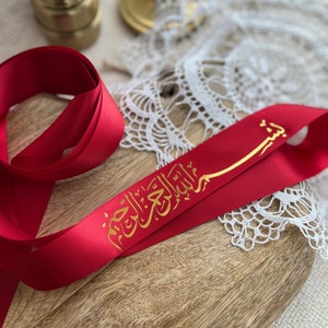 Peut inclure: Ruban de satin rouge avec calligraphie arabe dorée. Le ruban est posé sur une surface en bois, avec un napperon en dentelle blanche et un bougeoir doré en arrière-plan. Le ruban est d'un rouge vif.