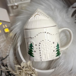 Op de afbeelding: Een witte keramische mok met een huisvormig deksel. De mok is versierd met groene kerstbomen, sterren en stippellijnen. De mok heeft een oor en staat op een wit, pluizig oppervlak.