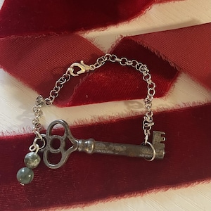 May include: A silver-toned key charm bracelet with a vintage aesthetic. The key is attached to a chain with a lobster clasp and two green and gray beads. The bracelet rests on a red velvet ribbon.