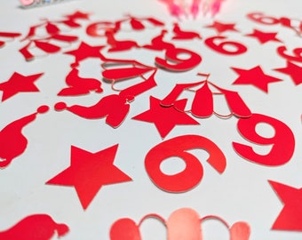 Puede incluir: Recortes de papel rojo, incluyendo estrellas, los números 6 y 9, y gorros de Papá Noel, están dispersos sobre una superficie blanca. Los recortes son brillantes y reflejan la luz, creando un ambiente festivo. La imagen está tomada desde un ángulo ligeramente elevado.