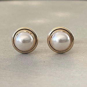 May include: A pair of gold-tone stud earrings. Each earring features a large, round, white pearl set within a gold bezel. The earrings are simple and elegant, suitable for everyday wear or special occasions.