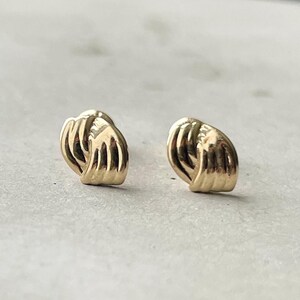 May include: A pair of gold-colored stud earrings. Each earring features a textured, knotted design. The earrings are displayed on a light gray surface.