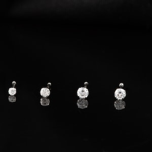 May include: Six silver nose rings with clear round gemstones of varying sizes. The rings are arranged in a row on a black background.